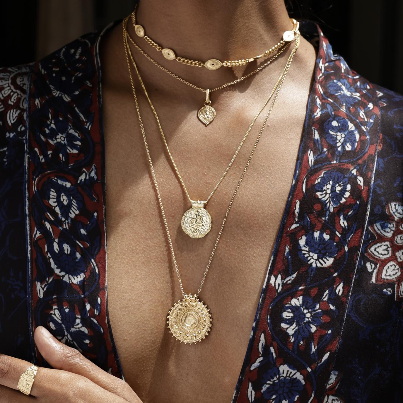 Close-up of a model wearing multiple gold necklaces with intricate designs, against a dark background. By Loft & Daughter
