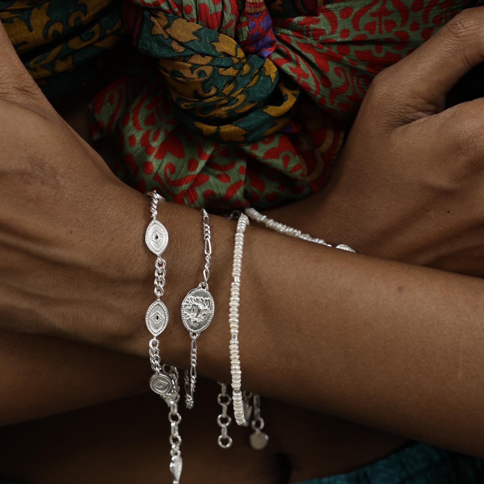 Close up of model wearing delicate silver stacking bracelets featuring evil eye charms and an oval Durga charm, worn with dainty pearl braclet.