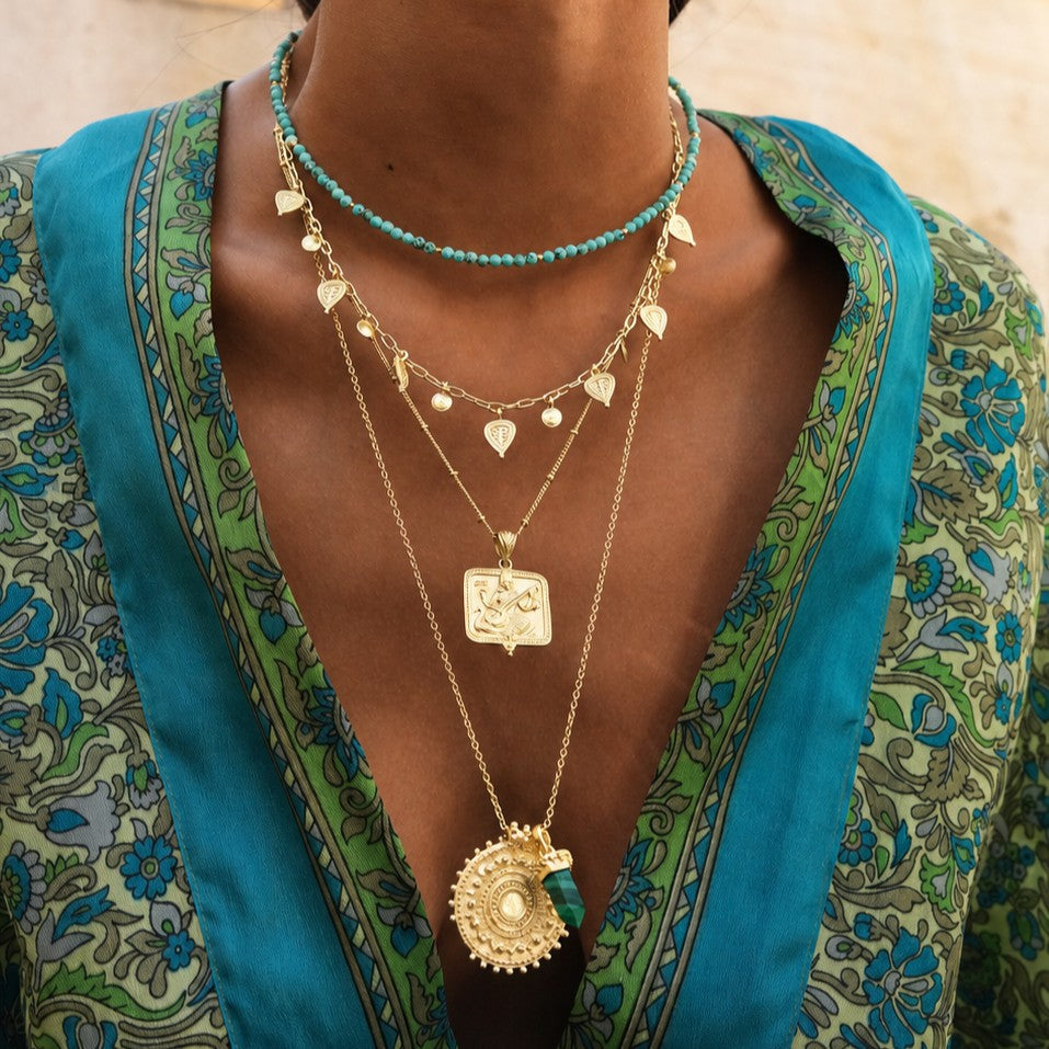 A close-up of a person wearing a square Saraswati Pendant. The necklaces include a short beaded choker, delicate chain layers with small gold charms, a rectangular pendant, and a larger circular pendant.