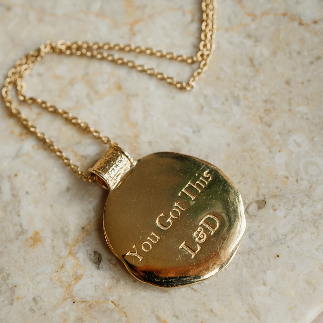 Gold necklace with a pendant engraved with 'You Got This L&D' on a marble surface.