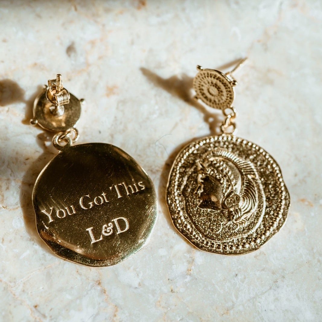 Gold earrings with 'You Got This' and initials on a marble surface