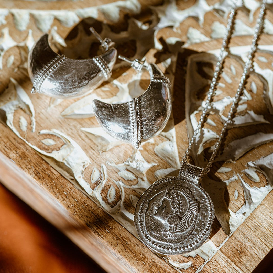 Silver You Got This Athena coin necklace on a textured wooden surface with domed hoop earrings