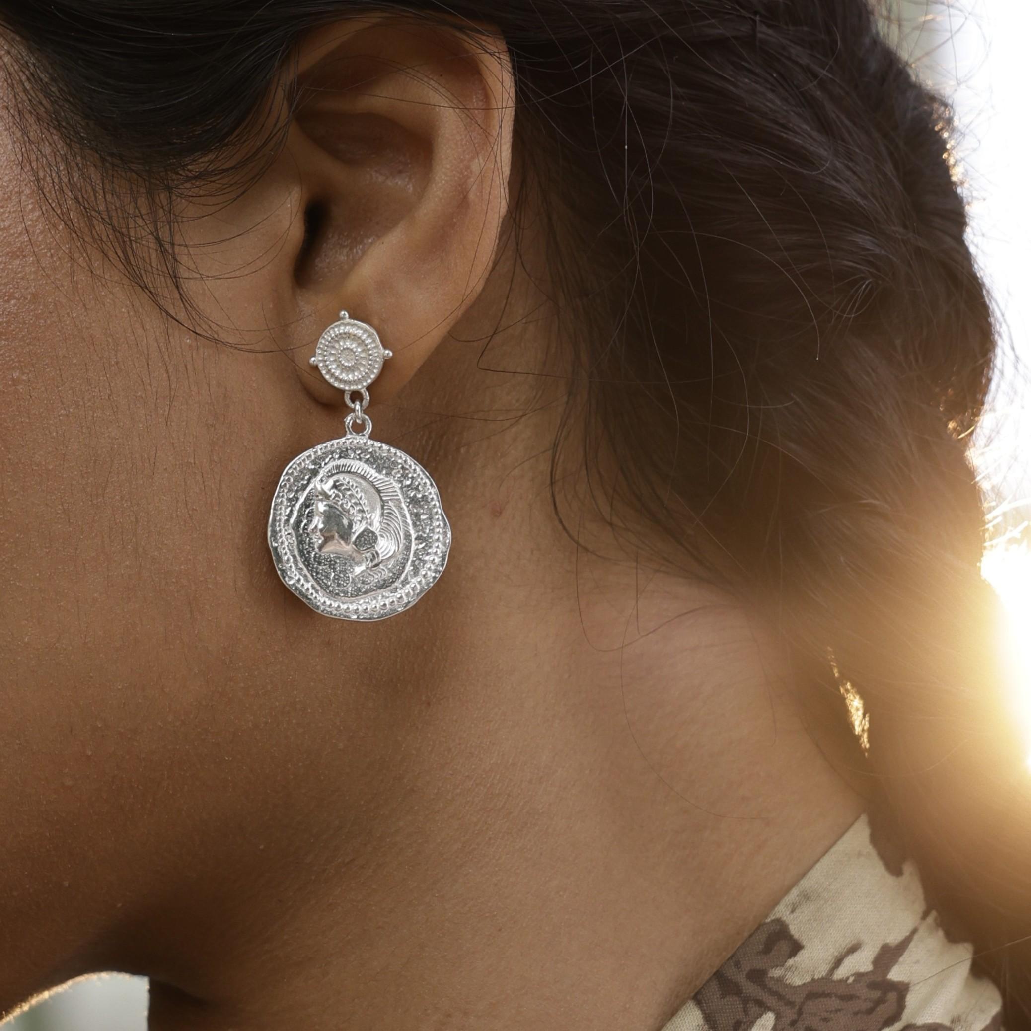 Close-up of a silver coin earring featuring goddess Athena worn by a model with a blurred background