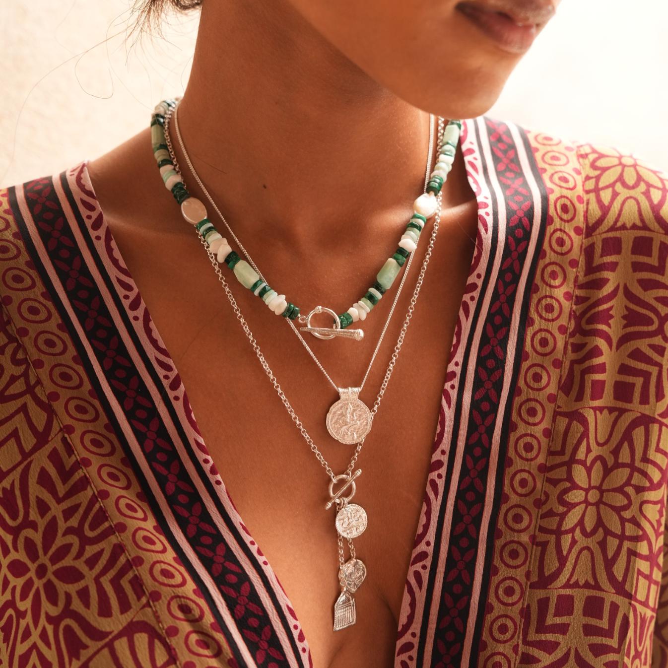 A close-up of a person wearing layered necklaces over a patterned bohemian-style top. The necklaces include a beaded strand with green and white beads, a silver clasp, and a silver coin pendant necklace.