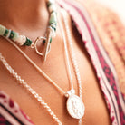 Close-up of layered necklaces worn on a person's neck, including a silver pendant necklace and a beaded necklace with green and white beads and a toggle clasp, against a patterned garment.