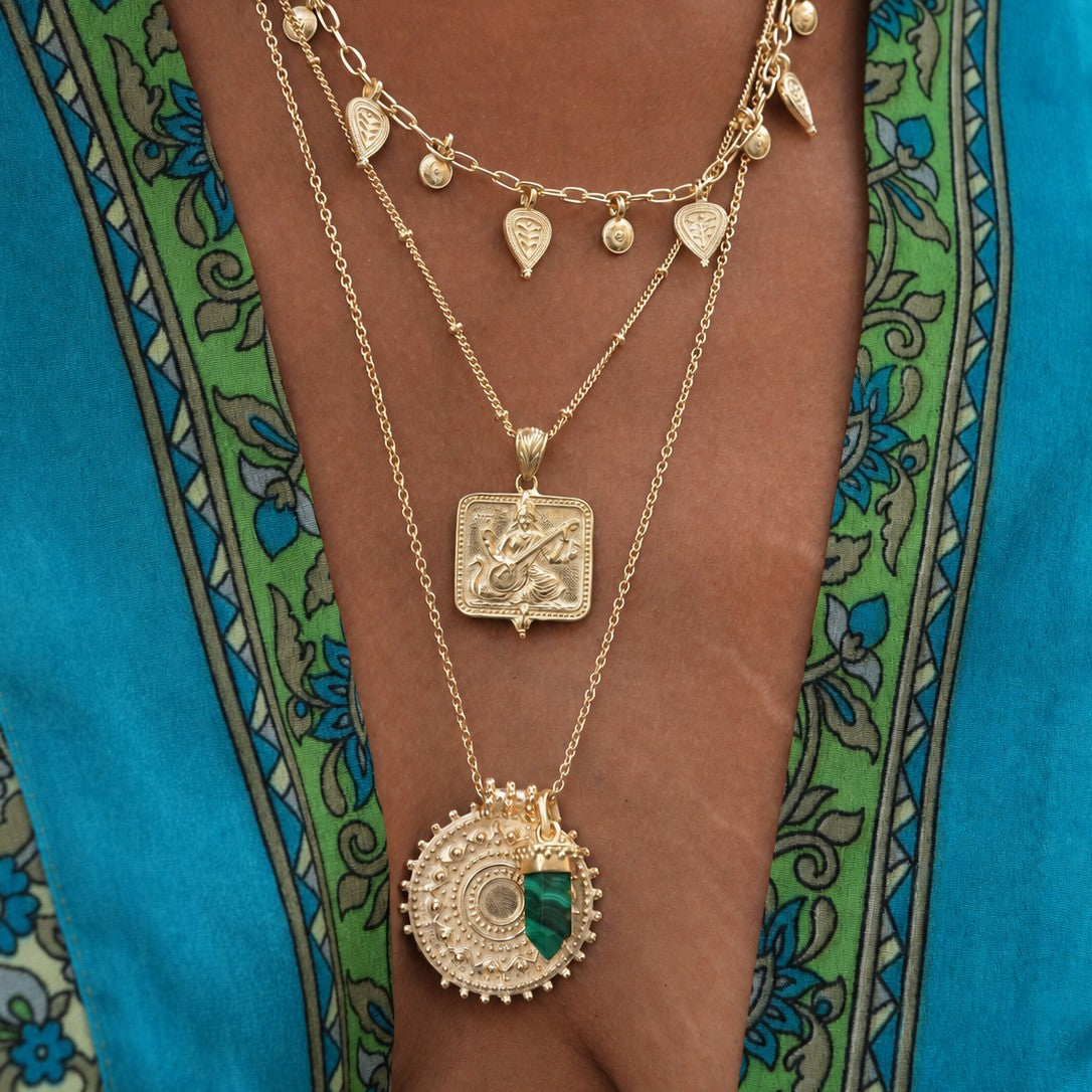 A close-up of a person wearing layered gold necklaces, including a circular pendant with a green stone accent, a square pendant, and smaller dangling charms, over a turquoise patterned garment.