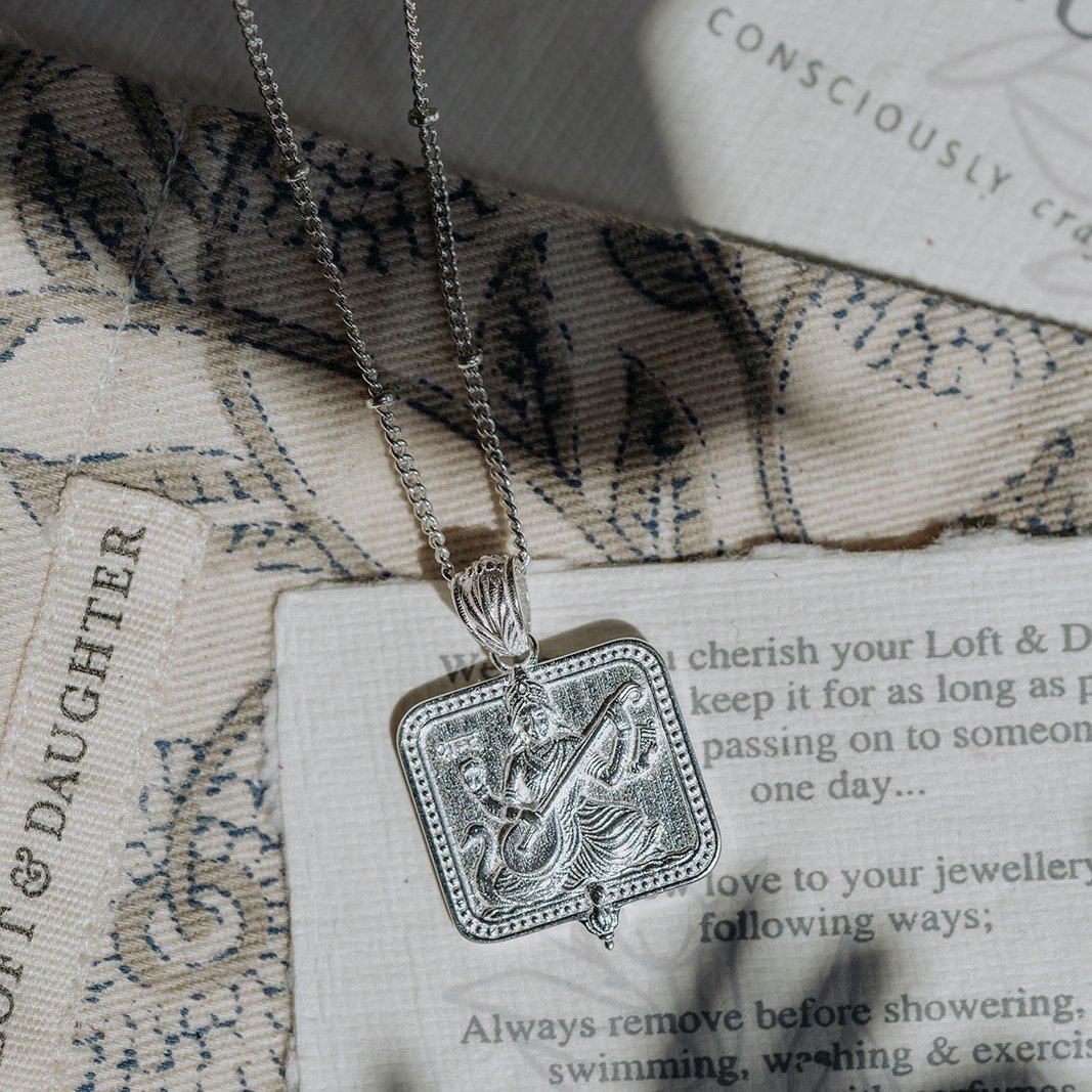 Silver Saraswati pendant necklace on a jewellery care card with woven fabric background